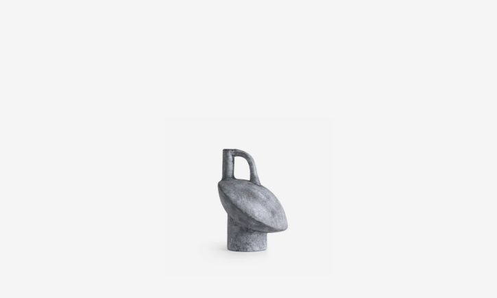A minimalist, abstract sculpture in grey featuring a small handle-like protrusion on top.